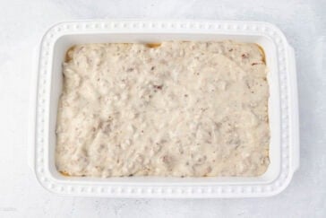A white rectangular baking dish filled with a thick, creamy white sauce mixed with ground meat. The dish is placed on a light-colored surface, showcasing the textured topping evenly spread across the top.