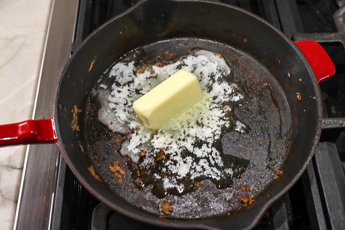 A red-handled cast iron skillet with a partially melted stick of butter in the center of the pan. The skillet has remnants of previously cooked food stuck to the surface, and is placed on a stove.