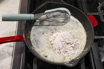 A red-handled cast-iron skillet on a stove contains a bubbling mixture of melted butter and flour, ready to be whisked into a roux. A green-handled whisk rests on the skillet edge, ready to combine the ingredients. Marble countertop visible on the side.