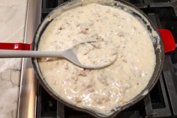 A red skillet on a stove contains a creamy sausage gravy. A gray ladle rests in the skillet, partially submerged in the thick, beige mixture with visible sausage bits. The stove and part of a marble countertop are visible in the background.
