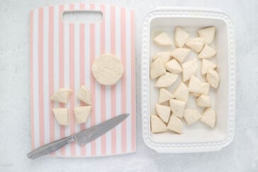 A pink and white striped cutting board with dough pieces and a knife on it is next to a white rectangular dish filled with triangular dough pieces. One round piece of dough is on the cutting board. The background is a light gray surface.