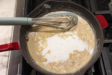 A whisk is resting in a red-handled skillet on a stove. The skillet contains a bubbling mixture of melted butter, flour, and milk, being prepared to make a creamy sauce or roux. The mixture is lightly browned and partially blended.