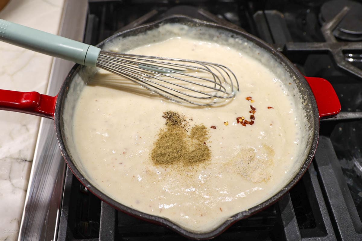 A whisk rests in a skillet filled with a creamy white sauce. The sauce contains visible specks of spices, including a pile of ground pepper and red pepper flakes, being mixed on a stove. The skillet has a red handle.