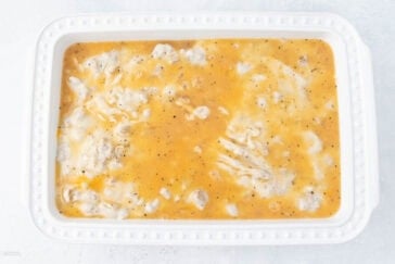 A white rectangular baking dish filled with an uncooked mixture of a yellowish cheesy sauce and small chunks of chicken. The dish is on a light-colored countertop. The surface of the mixture appears creamy with specks of black pepper.