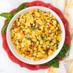 A vibrant corn salad sits in a white bowl with a red scalloped edge. The salad includes corn kernels, diced red bell peppers, and finely chopped herbs. Surrounding the bowl are basil leaves and a yellow-checkered napkin. The presentation is fresh and colorful.