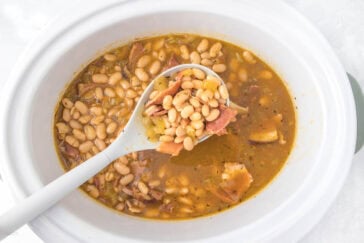 A white crockpot filled with cooked white beans and pieces of ham in a thick broth. A white ladle is lifting a portion of the beans and ham. The dish appears well-seasoned and hearty.