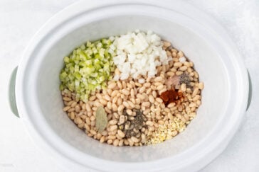 A slow cooker filled with uncooked white beans, chopped onions, diced celery, minced garlic, a bay leaf, and various ground spices. The ingredients are arranged in separate clusters over a white background.