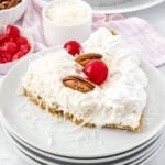 A slice of creamy white pie topped with three red cherries, pecans, whipped cream, and shredded coconut. The pie rests on a stack of three white plates. In the background, there are a small bowl of shredded coconut and a dish of red cherries.