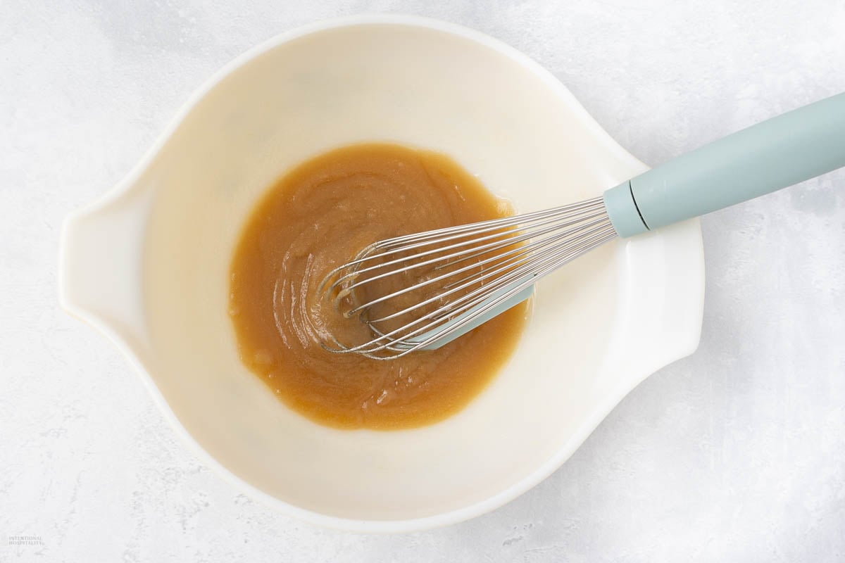 A white mixing bowl containing a thick, light brown mixture is shown from above on a light textured surface. A light blue-handled whisk is resting inside the bowl, partially submerged in the mixture.