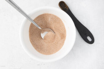 A white bowl containing a mix of cinnamon and sugar, with a spoon placed inside, rests on a light surface. Beside the bowl lies a black measuring spoon marked "1/2 tsp.