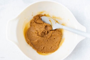 A white mixing bowl containing a brown dough-like mixture, with a gray spatula resting in the bowl. The contents appear to be partially mixed. The bowl is placed on a light-colored surface.