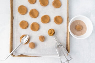 A baking tray lined with parchment paper holds several unbaked cookies, partially flattened and spaced apart. A bowl with a ball of cookie dough coated in cinnamon sugar is next to the tray, along with a cookie scoop and a spoon used for scooping and shaping the dough.