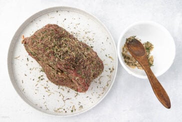 A raw, seasoned beef roast sits on a white plate. Next to it is a small white bowl with a wooden spoon resting inside, containing a mixture of herbs and spices. The background is a light, textured surface.