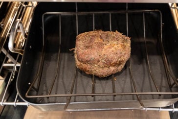 A seasoned roast beef is cooking in an oven, placed on a rack inside a roasting pan. The meat is browned and sprinkled with herbs. The oven rack and walls are visible. The surrounding area of the pan is clean and tidy.