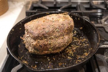 A seasoned roast beef sits in a black cast-iron skillet on a stovetop. The meat is browned evenly and covered with herbs, giving it a flavorful appearance. The stovetop burners are visible in the background.
