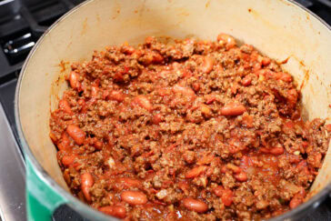 A pot of chili sits on a stovetop. The chili is a rich, hearty mixture of ground meat, red beans, and tomatoes in a thick sauce, seasoned with visible spices. There are bits of onion and garlic throughout the mixture.
