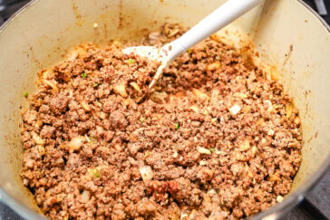A pot of seasoned ground beef mixed with bits of onions and spices being stirred with a white spoon. The mixture is sizzling and looks ready to be used as a filling or topping in various dishes.