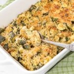 A white rectangular baking dish filled with a golden-brown zucchini casserole topped with breadcrumbs. A silver spoon is scooping out a portion, revealing the tender, baked zucchini underneath. The dish rests on a green and white checkered cloth.