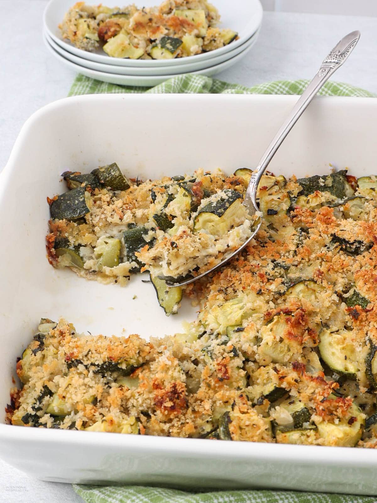 A white baking dish filled with a zucchini gratin topped with a golden, crispy breadcrumb crust. A serving spoon is placed in the dish, and a small portion has been served onto a plate in the background. A green cloth is partially visible underneath the baking dish.