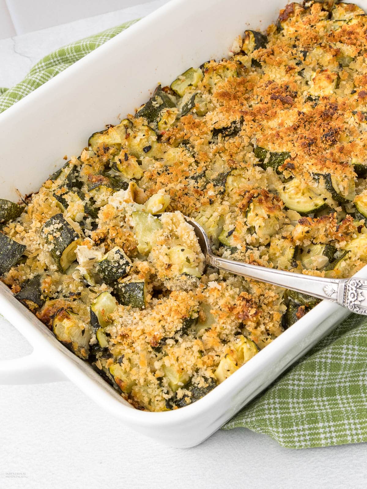 A white casserole dish filled with baked zucchini gratin topped with golden breadcrumbs. A silver serving spoon rests inside the dish, suggesting that some portions have already been taken. A green and white checkered cloth is partially visible underneath the dish.