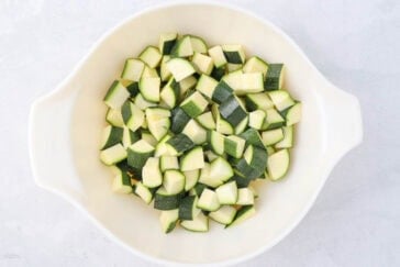 Chopped zucchini pieces in a white mixing bowl on a light gray background. The zucchini is cut into small, uneven chunks with a mix of green skin and pale flesh visible.