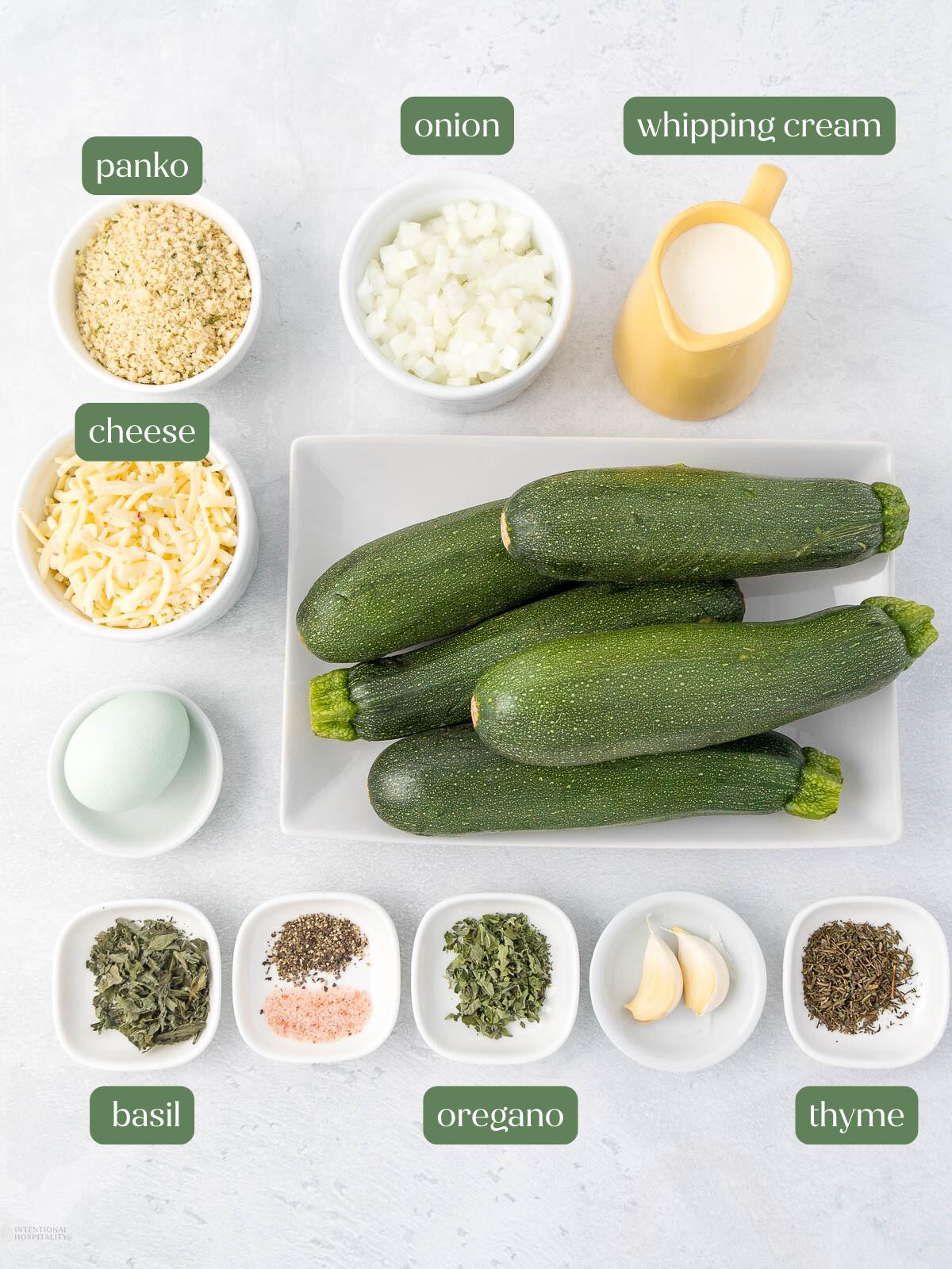 Ingredients for a zucchini recipe are shown, including panko breadcrumbs, shredded cheese, a whole egg, chopped onions, whipping cream, whole zucchinis, minced basil, salt and pepper, dried oregano, fresh thyme, and garlic cloves, all arranged on a white surface.