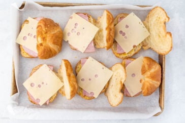 Tray of four croissant sandwiches with ham and slices of Swiss cheese, placed on parchment paper. Some croissants are open, revealing the ingredients inside.