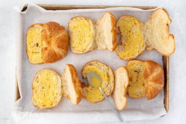 Slices of croissants arranged on a baking tray lined with parchment paper. A spoon with a yellow substance, possibly butter or custard, rests among the slices, suggesting a preparation or baking process.
