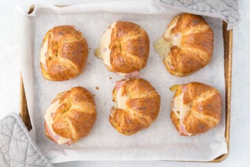 Freshly baked croissant sandwiches filled with ham and cheese, placed on a parchment-lined baking tray. Two gray oven mitts are visible on either side of the tray.
