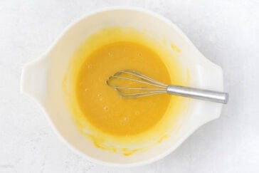 A white mixing bowl filled with a smooth, yellow batter. A metal whisk rests inside the bowl, partially submerged in the batter. The bowl is placed on a light gray countertop.