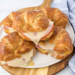 A wooden platter with croissant sandwiches filled with sliced ham and melted cheese. A striped blue and white napkin is partially visible on the side.