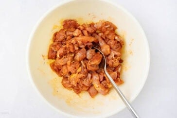 A bowl filled with marinated chicken pieces, seasoned with spices, and a spoon resting in it, set on a white background.