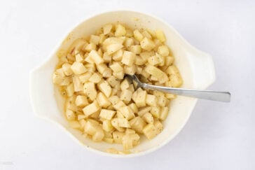 A white bowl filled with seasoned, cubed potatoes. A silver spoon rests inside the bowl on the right side. The bowl is placed on a white surface.