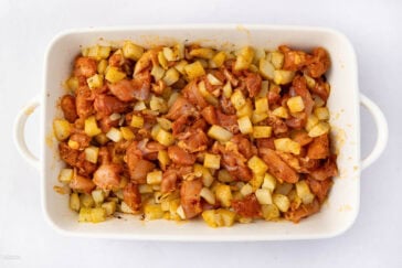 A white baking dish filled with seasoned diced potatoes and chunks of raw marinated chicken, ready for baking. The ingredients are well mixed and spread evenly across the dish.