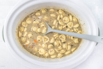 A white slow cooker filled with creamy chicken soup, featuring pasta, chunks of vegetables, and herbs. A gray spoon rests inside, ready for serving. The image captures both the texture and ingredients of the comforting dish.