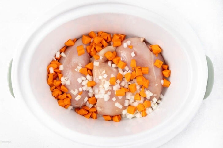 Raw chicken breasts, chopped carrots, and diced onions in a white oval crockpot on a white surface.