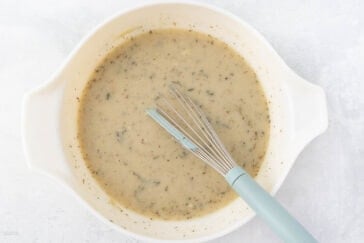A white bowl containing a creamy, herb-speckled sauce with a whisk resting inside. The sauce is light beige, indicating a blend of ingredients, and the background is a light gray surface.
