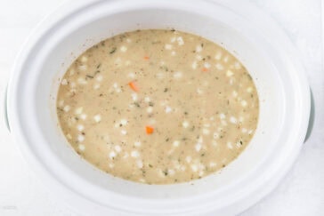 A slow cooker filled with a creamy soup containing diced potatoes and carrots, sprinkled with herbs. The soup appears thick and hearty, with visible chunks of vegetables. The white slow cooker is shown from a top view.