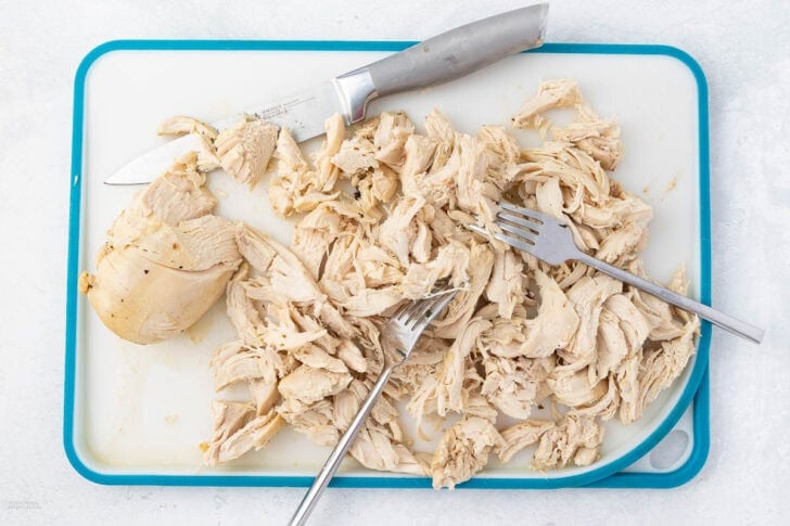 Shredded cooked chicken placed on a white and blue cutting board, accompanied by two forks used for shredding and a knife.