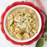 A bowl of creamy chicken tortellini soup topped with grated cheese and fresh herbs. The soup includes shredded chicken, tortellini pasta, and diced carrots, served in a round white bowl on a red plate. Fresh basil leaves and a napkin are beside the bowl.