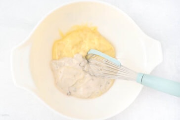 A white bowl contains two different types of sauce, one yellow and the other cream-colored with specks, being mixed together with a whisk.
