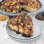 A slice of peanut butter pie on stacked white plates. The pie has a chocolate cookie crust, topped with chocolate, peanut butter cups, peanuts, and chocolate drizzle. In the background, the rest of the pie and ingredients are visible.