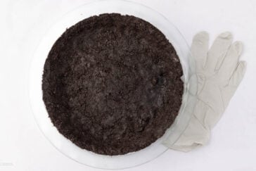A clear glass pie dish filled with dark soil sits on a white surface next to a pair of white rubber gloves.