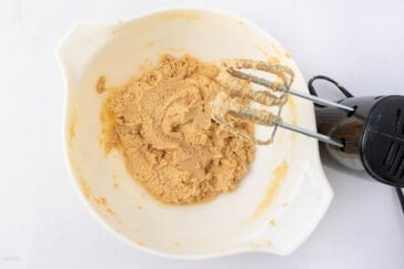 A white mixing bowl with creamy brown batter, alongside a black electric hand mixer resting inside. The bowl is placed on a white surface.