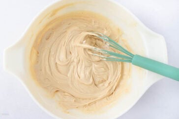A mixing bowl with creamy batter being whisked with a teal-colored whisk. The bowl is white, and the mixture is smooth and light brown, indicating it's been recently stirred.