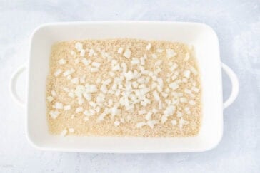 A white baking dish filled with uncooked rice and chopped onions is placed on a textured light-colored surface.