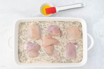 Raw chicken breasts in a white baking dish with a creamy herb marinade. A small bowl with a red silicone brush and oil is nearby on a light surface.