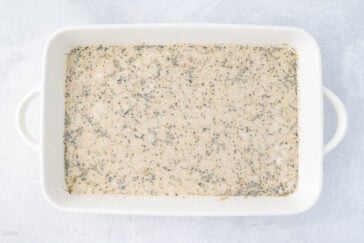 A rectangular white baking dish containing a creamy mixture with black specks is placed on a light gray background. The dish has handles on both sides.