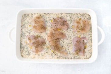 Raw chicken breasts seasoned with herbs and pepper in a creamy sauce, placed in a white baking dish.