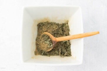 A wooden spoon rests in a square white bowl filled with dried herbs. The herbs are finely chopped, with a mix of green leaves and ground seasoning. The background is a light textured surface.